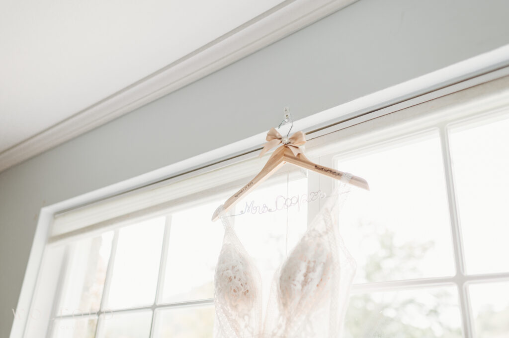 A delicate lace wedding gown hangs from a personalized wooden hanger engraved with 'Mrs. Cooper,' adorned with a soft beige ribbon. The gown is beautifully showcased in front of a bright window, allowing natural light to highlight its intricate lace detailing. A simple and elegant bridal moment captured in preparation for the wedding day