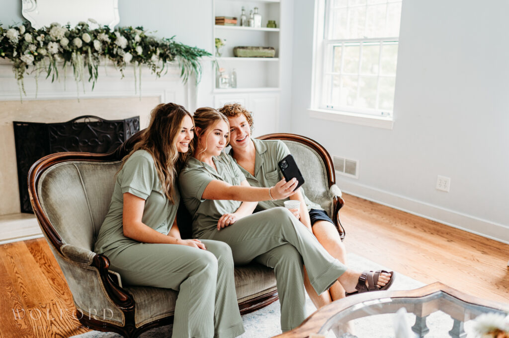 A joyful group of bridesmaids dressed in matching gowns gather closely, smiling and laughing as they pose for a selfie. The phone is held up by one of the bridesmaids while the others lean in, capturing the excitement and fun of the wedding day. Their vibrant energy and playful camaraderie radiate through the candid moment.