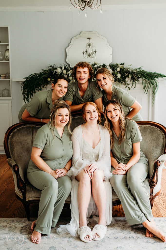 A group of bridesmaids pose together in matching pajamas, standing casually with playful expressions and relaxed postures. Some hold coffee mugs or champagne glasses, while others drape their arms around each other. The cozy setting reflects a fun and intimate moment shared during the wedding morning preparations, highlighting their close bond and excitement.