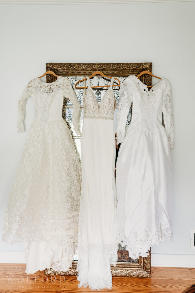 Three generations of wedding dresses displayed side by side: a vintage long-sleeved lace gown with intricate floral patterns, a modern sleeveless gown with a plunging neckline and delicate lace detailing, and a classic satin dress with lace sleeves and a full skirt. The dresses hang on wooden hangers against an ornate vintage mirror, symbolizing the timeless tradition of love and family.